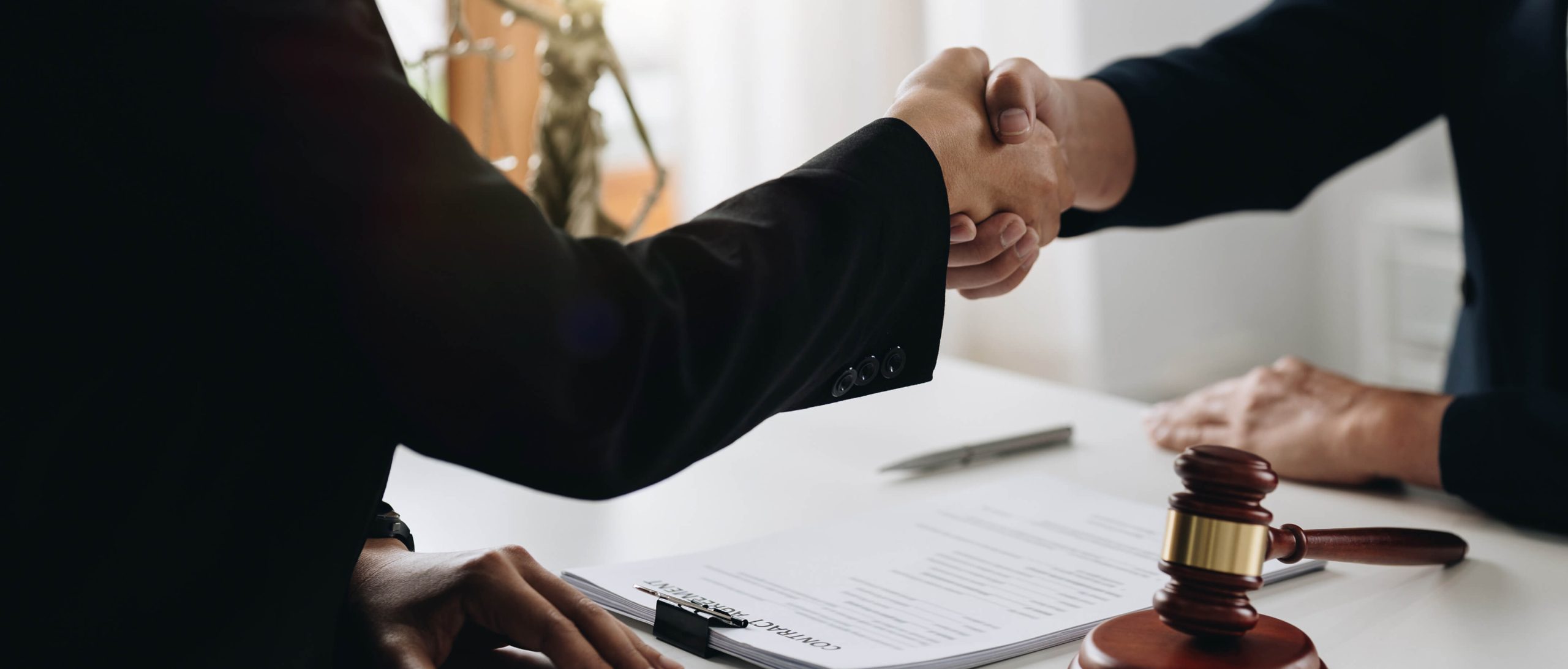 Two people in business attire shaking hands over a desk with legal documents, a pen, and a judge's gavel visible.