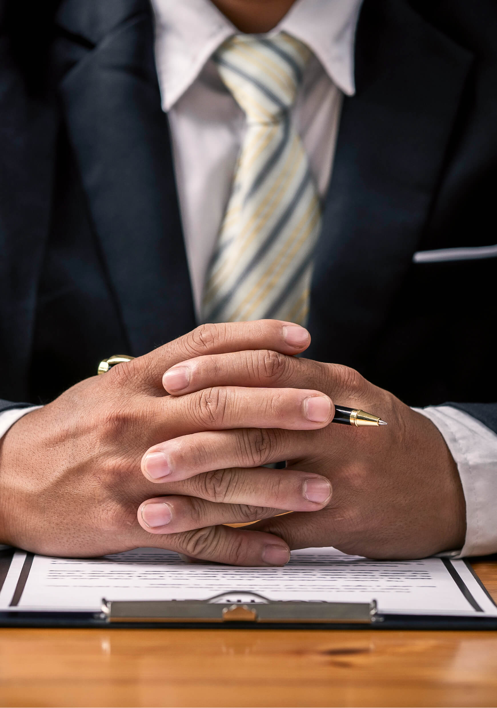 A person in a suit sits at a table with hands clasped, holding a pen, in front of a clipboard with documents.