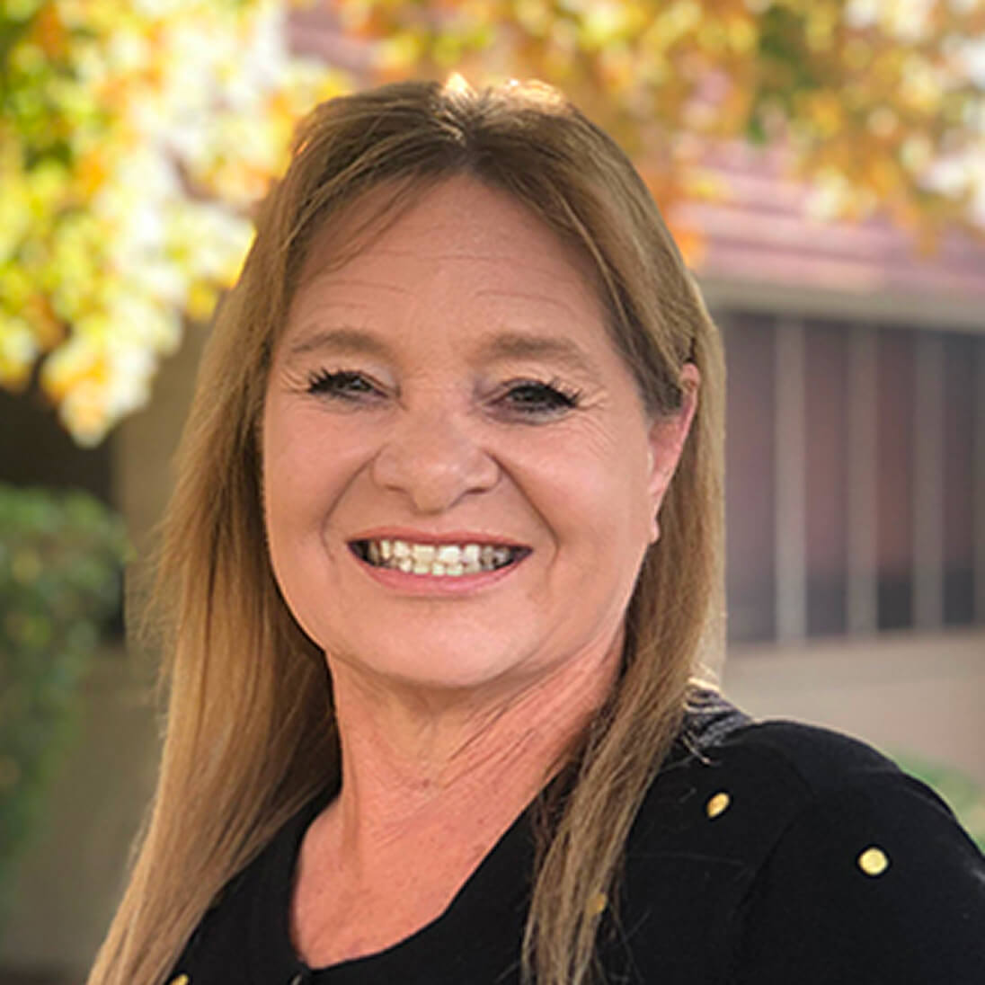Mujer sonriente con el pelo largo de color castaño claro y una blusa negra, de pie al aire libre con árboles y un edificio al fondo.