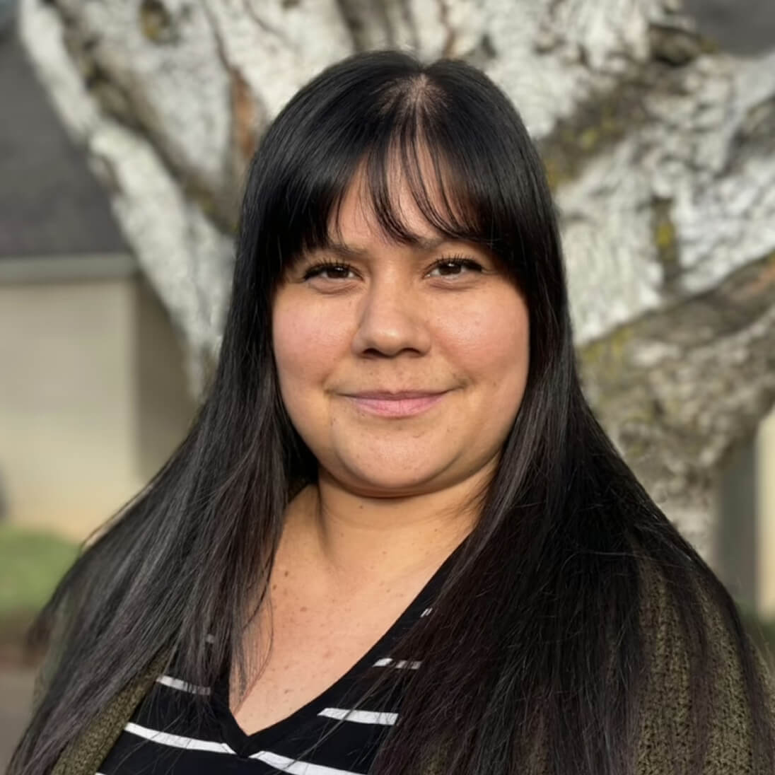 Una mujer con el pelo largo y oscuro y flequillo posa al aire libre frente a un árbol, vistiendo una camisa de rayas y un jersey verde.
