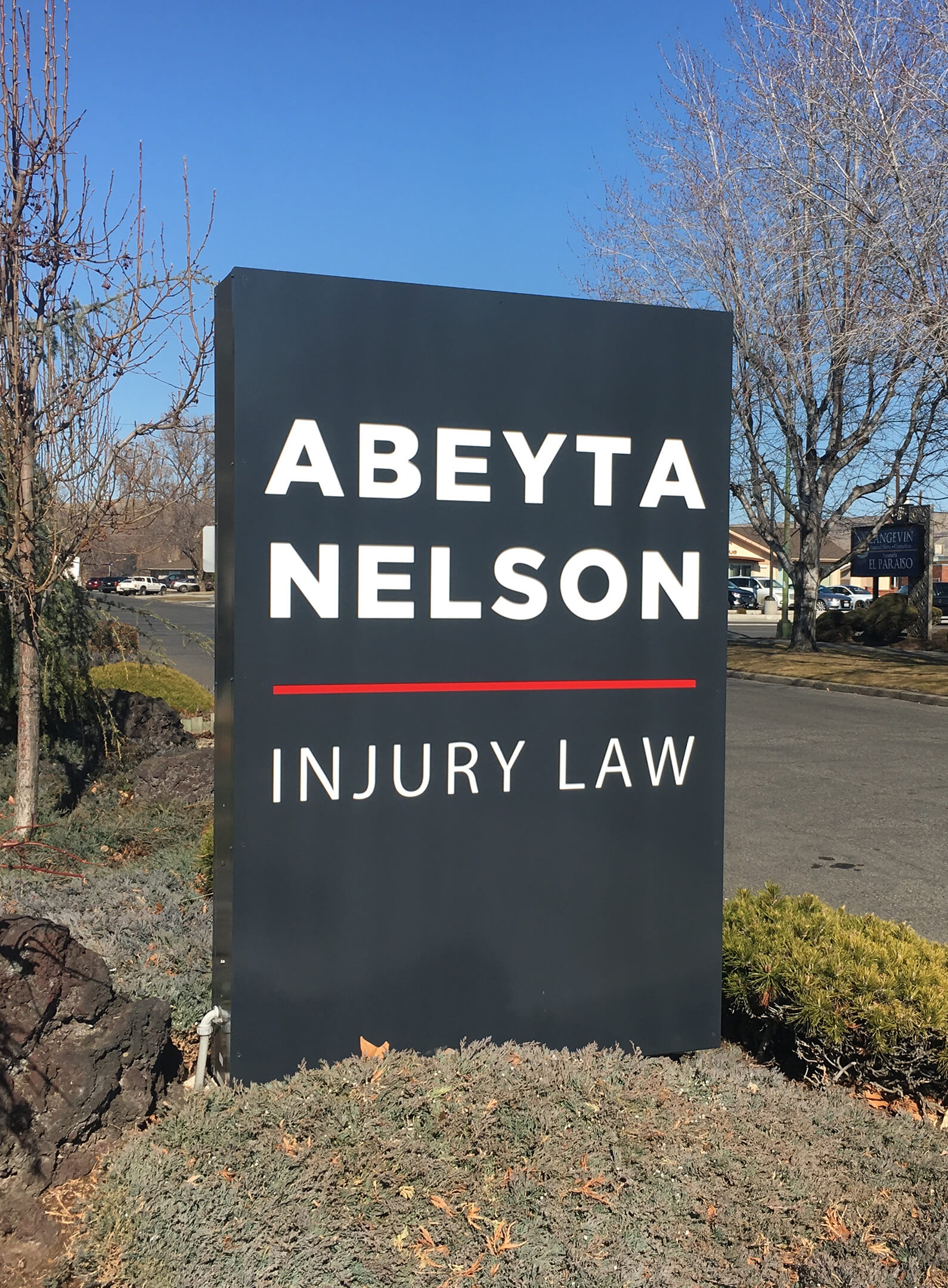 A large outdoor sign reads "Abeyta Nelson Injury Law" with white and red text on a black background, surrounded by shrubbery and trees.