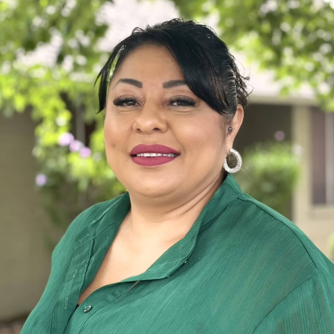 Una mujer de pelo oscuro con una blusa verde posa al aire libre, sonriendo, con árboles y un edificio al fondo.