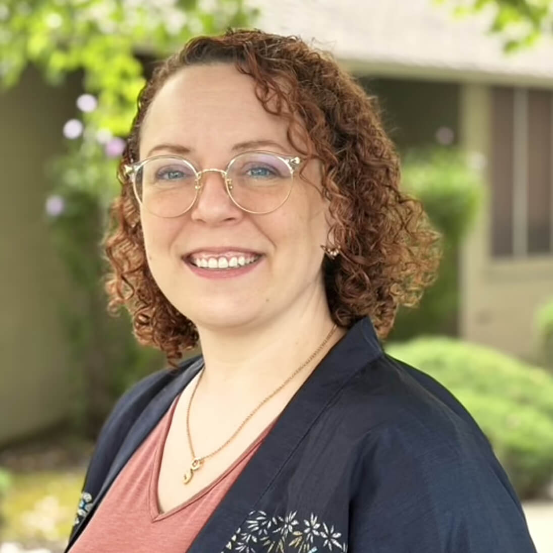 Una mujer con el pelo rizado, gafas y una chaqueta azul posa al aire libre, sonriendo a la cámara. Se ve vegetación y un edificio al fondo.