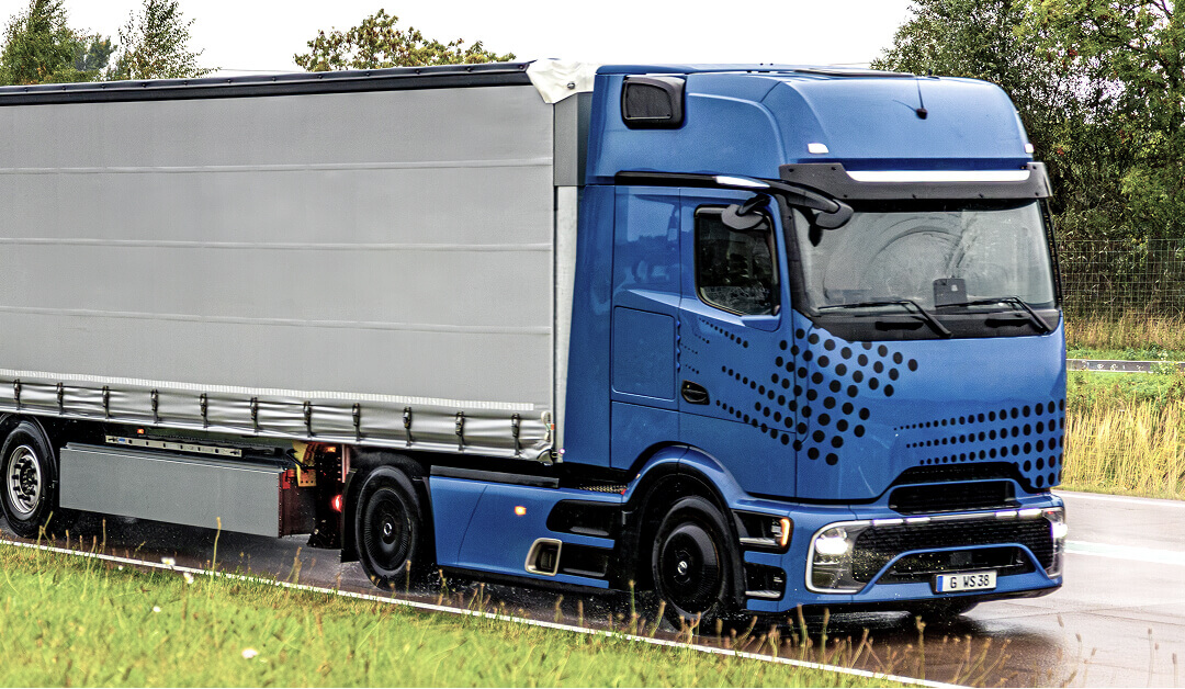 Un tráiler azul con remolque cubierto circula por una carretera mojada, rodeado de hierba y árboles.