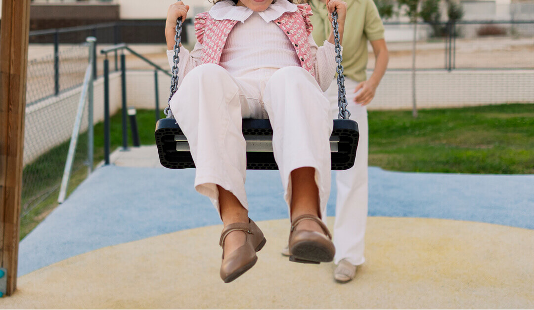 Un niño con ropa blanca está siendo empujado en un columpio por un adulto en un patio de recreo.