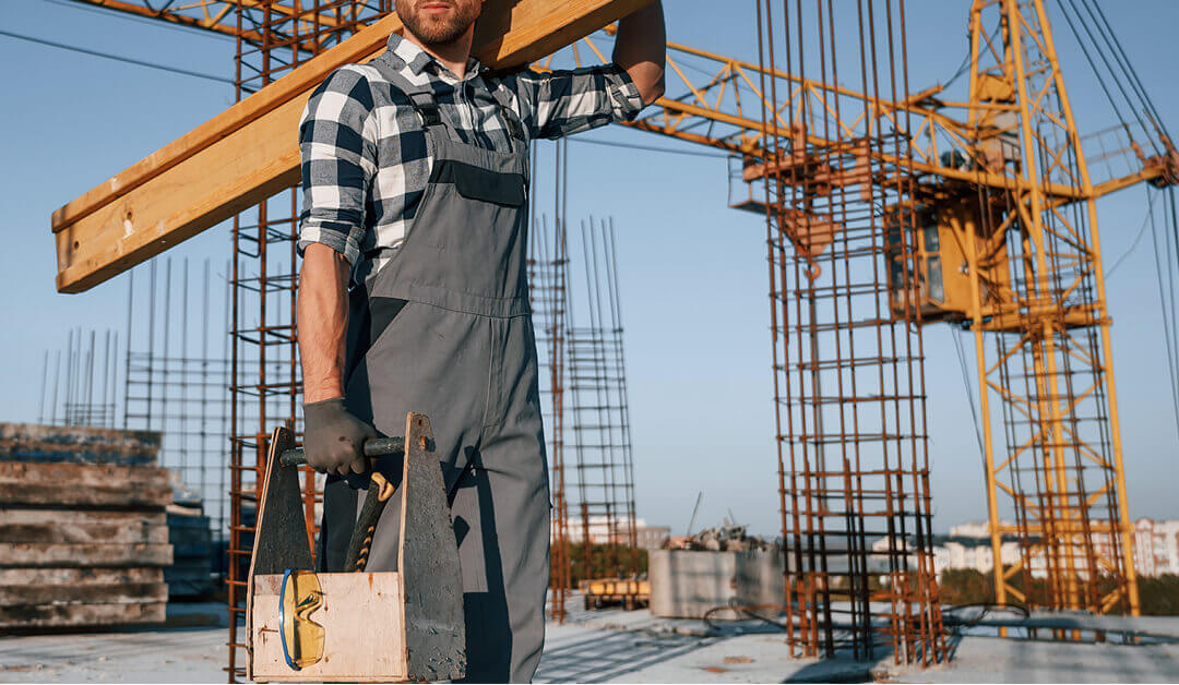 Un trabajador de la construcción con mono lleva una viga de madera al hombro y sostiene una caja de herramientas, con varillas metálicas y una grúa al fondo en una obra.