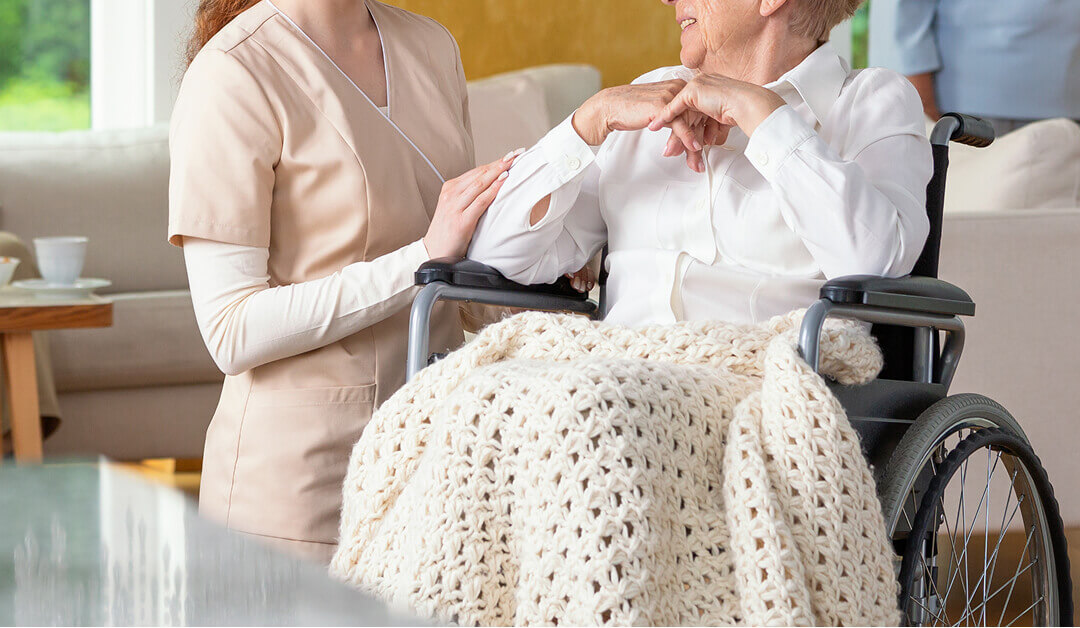 Un cuidador está de pie junto a una mujer mayor en silla de ruedas cubierta con una manta tejida, sosteniendo suavemente su brazo en un entorno doméstico.