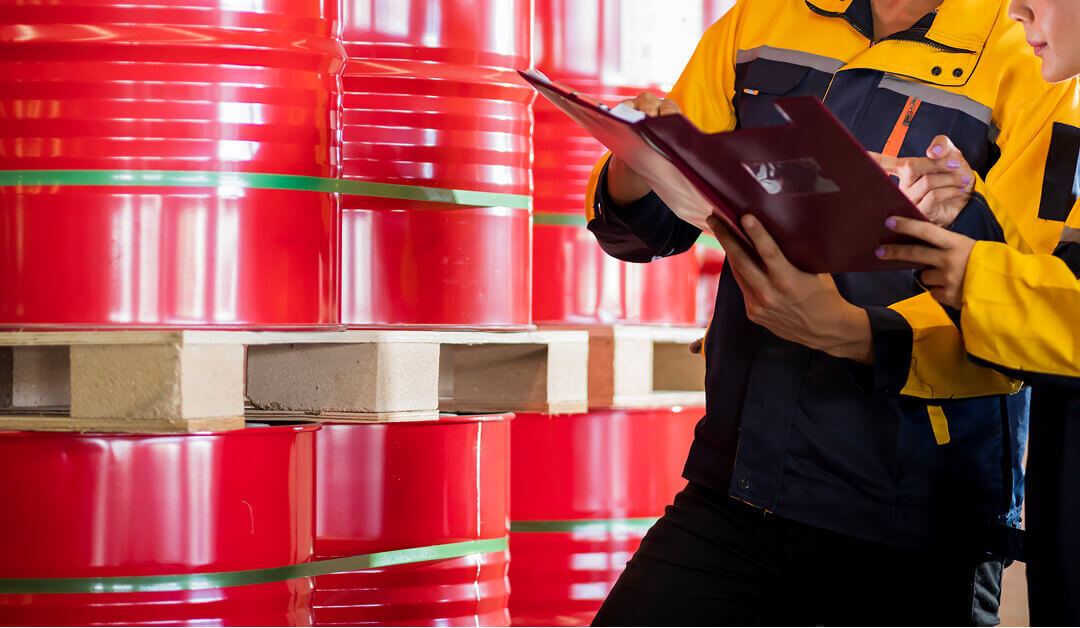 Dos trabajadores con uniformes amarillos y azul marino inspeccionan y registran información en un portapapeles junto a barriles rojos apilados sobre palés de madera.