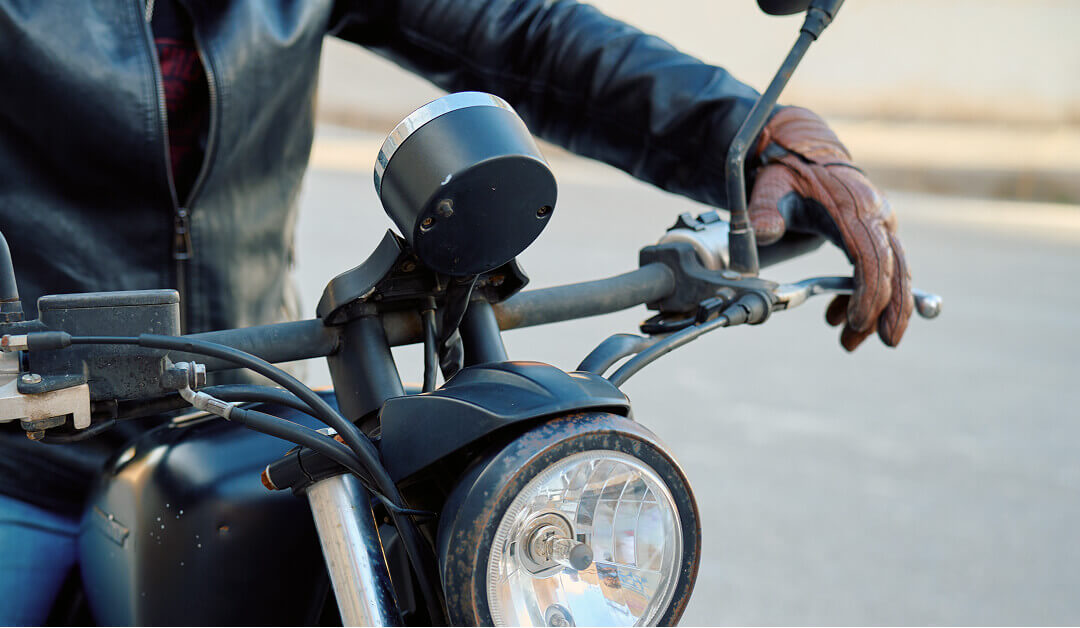 Persona con chaqueta de cuero y guantes marrones sujetando el manillar de una motocicleta negra, vista desde el frente.