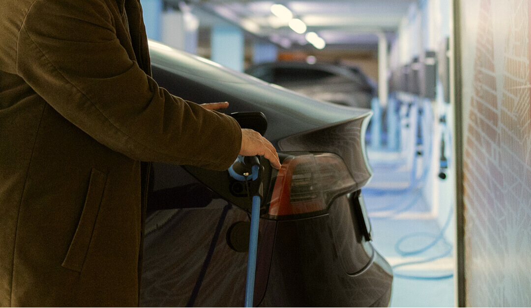 Una persona con un abrigo marrón enchufa un cable de carga en la parte trasera de un coche eléctrico negro en una estación de carga interior.