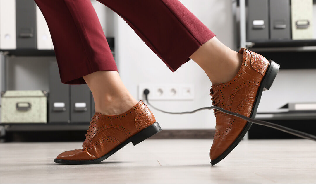 Persona con zapatos de vestir marrones y pantalones color burdeos tropezando con un cable eléctrico negro en el suelo de una oficina con estanterías y enchufes al fondo.