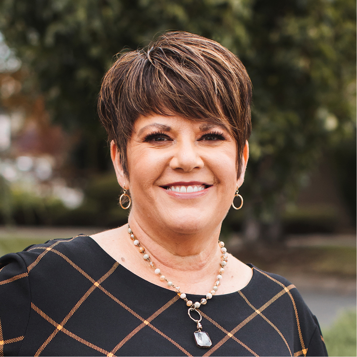 Una mujer con el pelo castaño corto, que viste una blusa negra estampada, pendientes dorados y un collar de cuentas, sonríe al aire libre con vegetación al fondo.