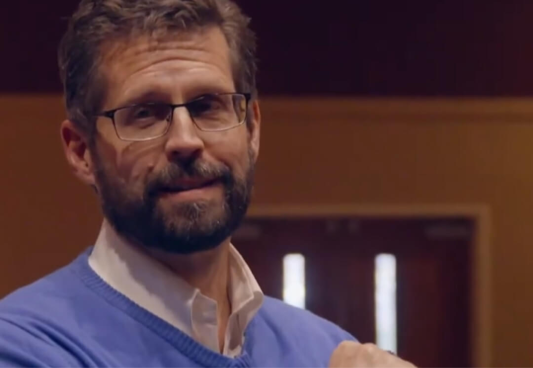 A man with glasses, a beard, and short hair wearing a blue sweater and white shirt indoors, standing in front of double doors.