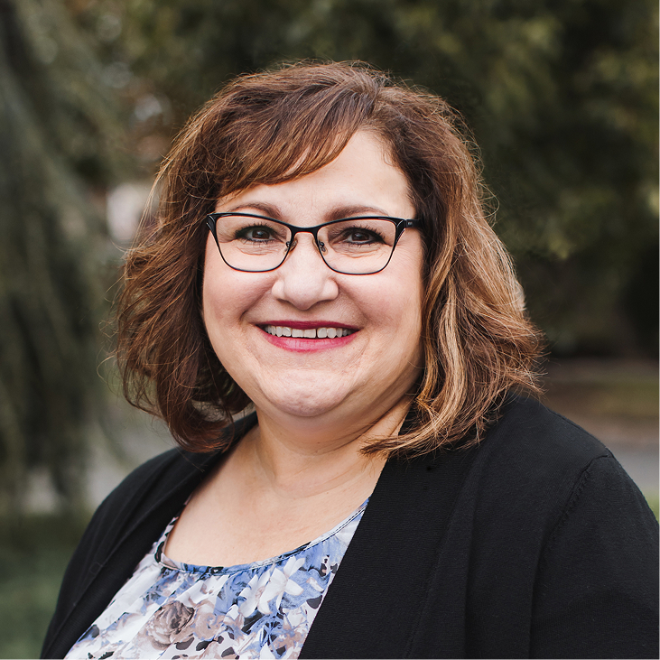 Una mujer con el pelo castaño hasta los hombros y gafas sonríe al aire libre, vistiendo una chaqueta negra sobre una blusa estampada. Se ven árboles desenfocados al fondo.