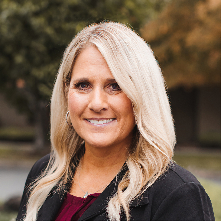 Una mujer con el pelo largo y rubio, que viste una americana oscura y una blusa granate, posa al aire libre sonriendo a la cámara con árboles desenfocados al fondo.